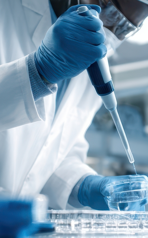Eine Person im Labor trägt Schutzbrille und blaue Handschuhe und pipettiert eine klare Flüssigkeit in eine Petrischale. Daneben stehen mehrere leere Reagenzgläser in einem Rack.