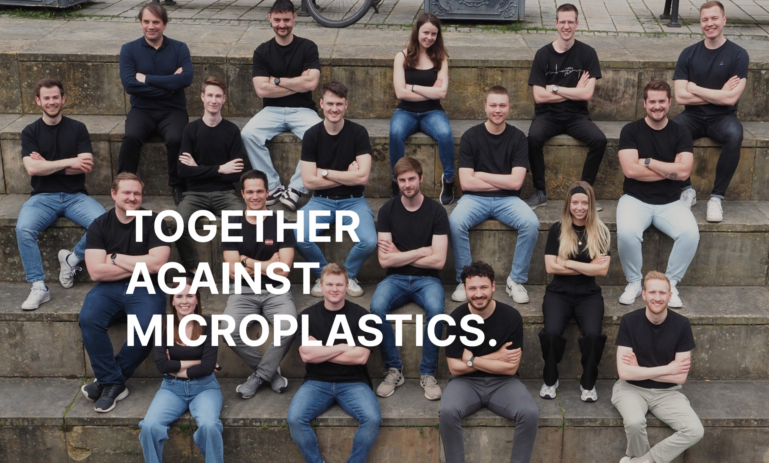 Alle Mitarbeitenden sitzen auf einer Steintreppe mit verschränkten Armen und schauen in die Kamera, die Über ihnen ist. Darüber liegt der Schriftzug: Gemeinsam gegen Mikroplastik.
